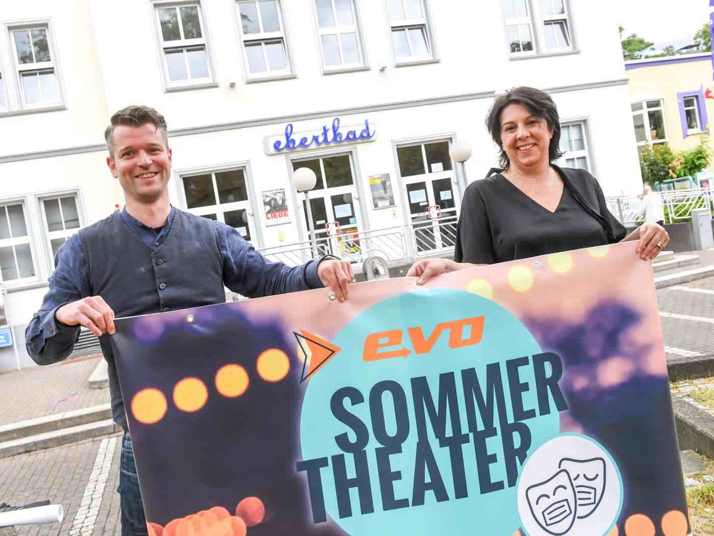 Zwei Personen stehen vor dem Ebertbad und halten ein Werbeplakat für das evo Sommertheater hoch.