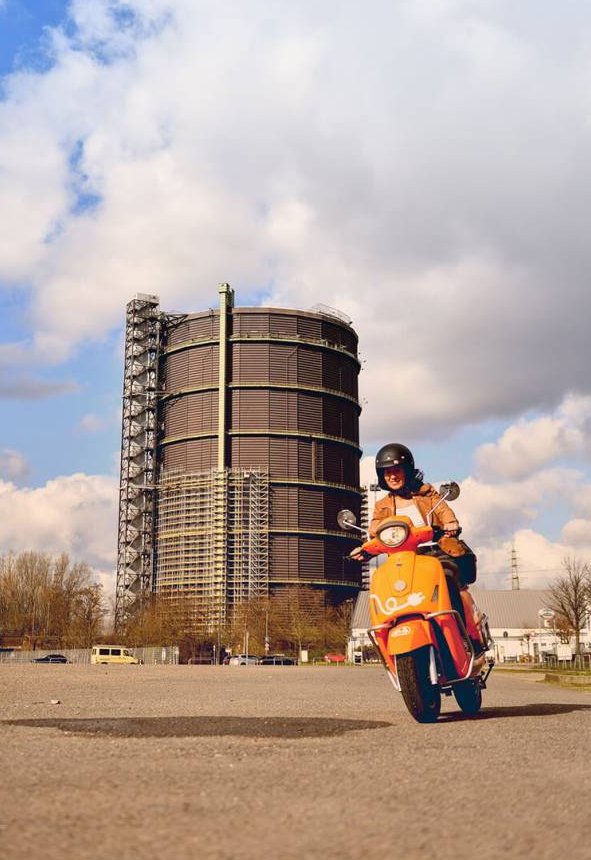 Eine Frau fährt mit einem evo Roller. Im Hintergrund ist das Gasometer zu sehen.