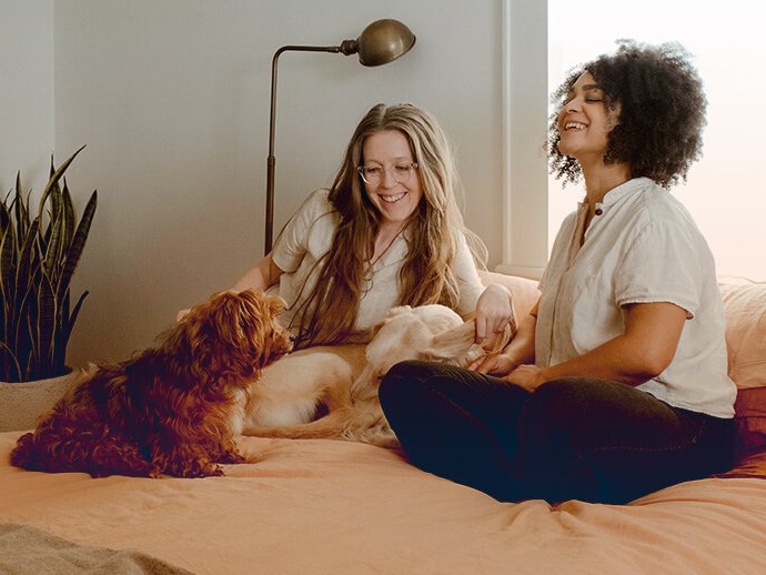 Zwei Frauen sitzen lachend auf einem Bett. Sie streicheln zwei Hunde, die mit ihnen auf dem Bett sind.