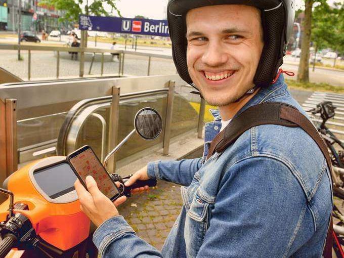 Ein junger Mann sitzt grinsend auf einem evo Roller und hat die Sharing-App auf dem Handy geöffnet.