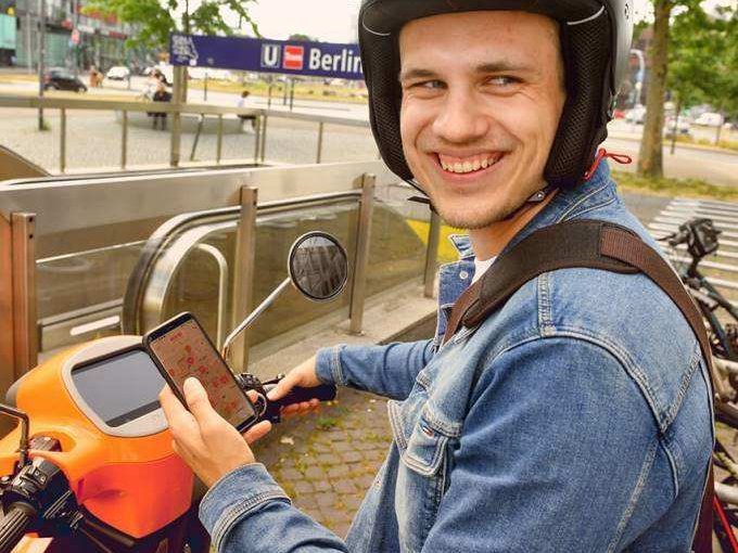 Ein junger Mann sitzt grinsend auf einem evo Roller und hat die Sharing-App auf dem Handy geöffnet.