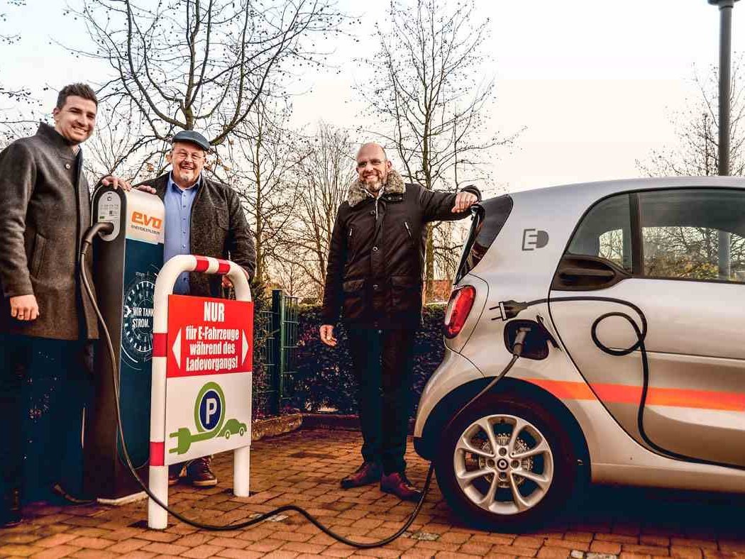 Drei Männer posieren neben einer Ladestation, an der gerade ein E-Auto lädt.