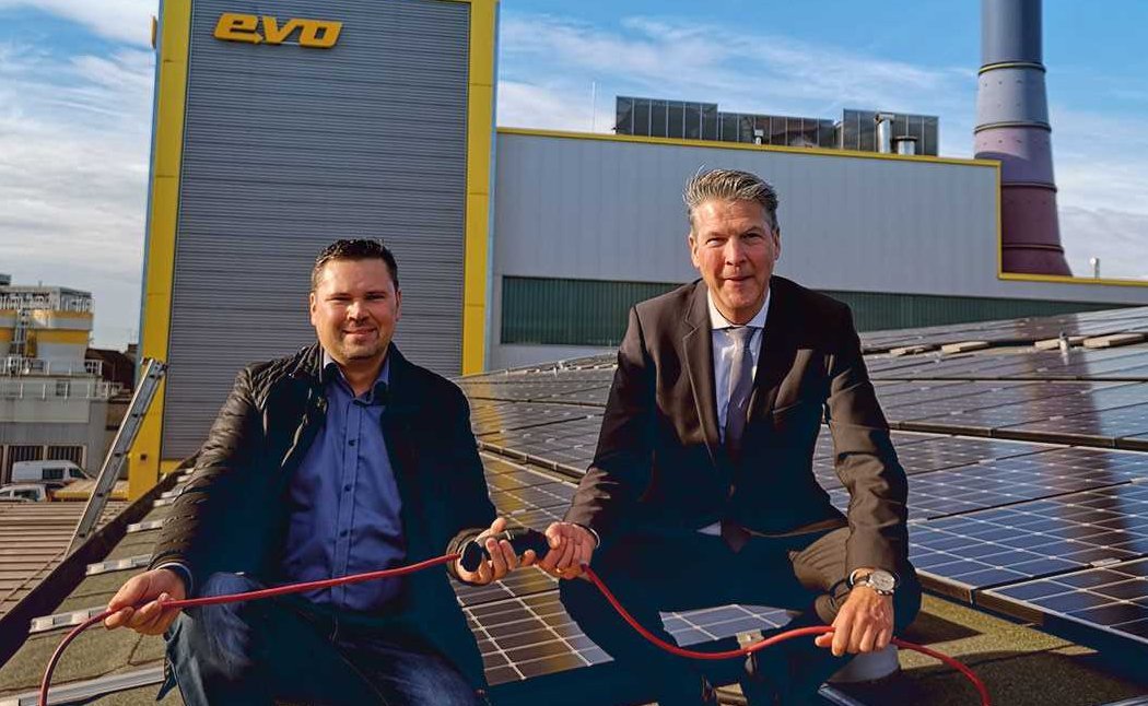 Zwei Männer sitzen zwischen Solarpanels auf einem Dach, vor dem evo Kraftwerk.