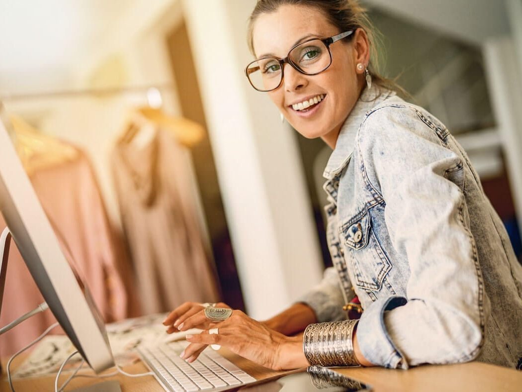 Eine Frau sitzt vor ihrem Computer und lächelt in die Kamera.