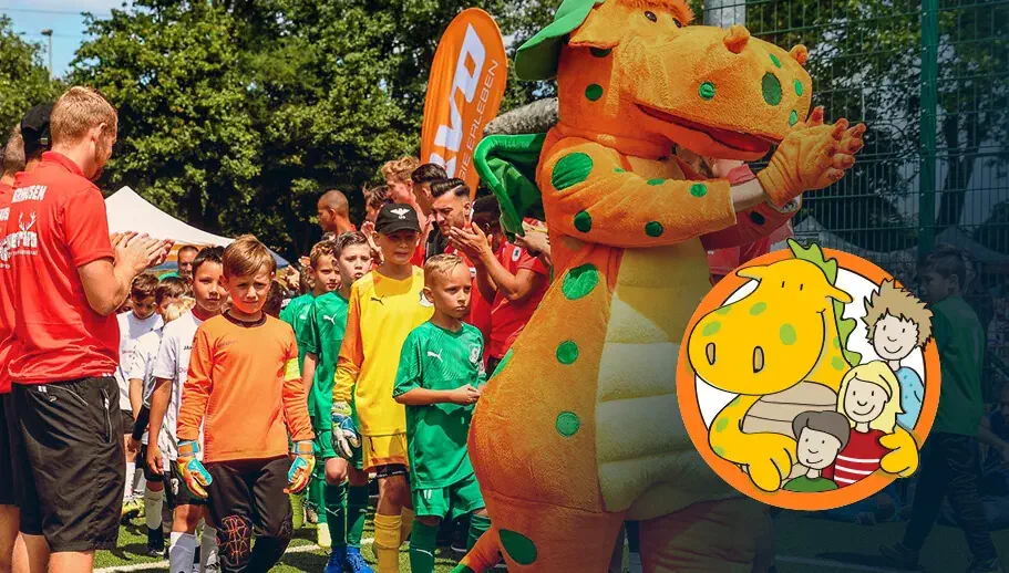 Eine Gruppe von Kindern in Fußballtrikots laufen in einer Reihe richtung Fußballplatz. Um sie herum stehen mehrere Spieler, die ihnen applaudieren. Ganz vorne in der Reihe läuft unser Rasmus-Maskottchen.