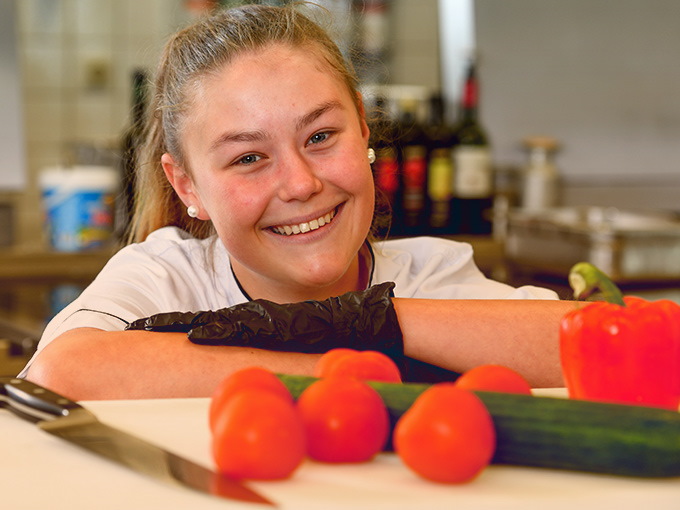 Eine junge Köchin, die lächelnd mit Gurken und ein paar Tomaten posiert.