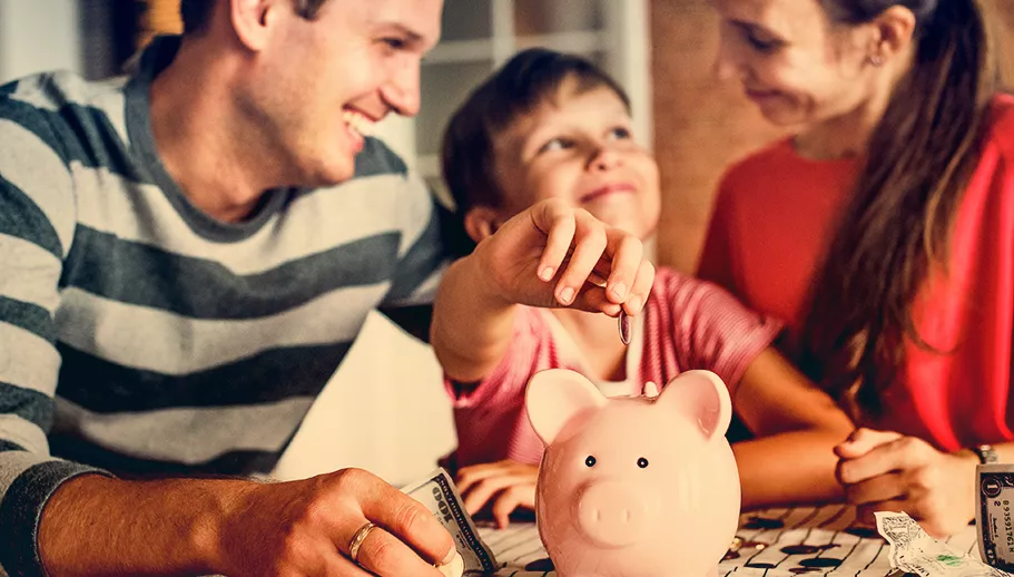 Eine Familie ist zu sehen. Der Sohn steckt gerade eine Münze in ein Sparschwein.