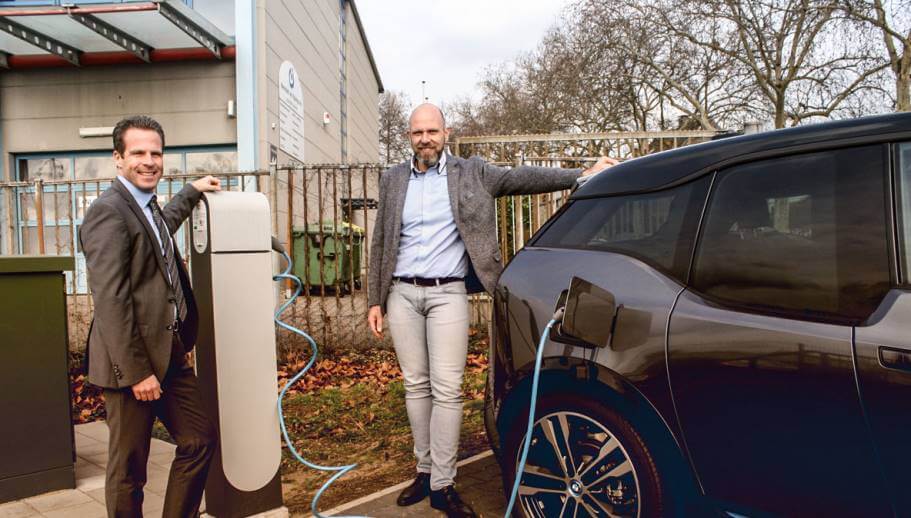 Zwei Männer posieren vor einem Elektroauto, welches gerade mit einer Ladestation verbunden ist. 