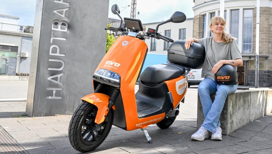 Eine junge Frau sitzt vor dem Hauptbahnhof Oberhausen. Neben ihr steht ein evo-Roller. Sie hat einen Helm auf dem Schoß.