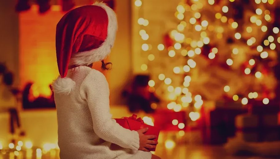 Ein kleines Kinde, welches ein Geschenk in der Hand hält und vor einem Weihnachtsbaum sitzt.